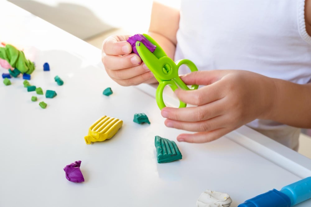 The Importance of Scissor Skills in Child Development: Tips and Activities to Get Started 7 Child using plastic safety scissors to cut play dough to help build scissor skills